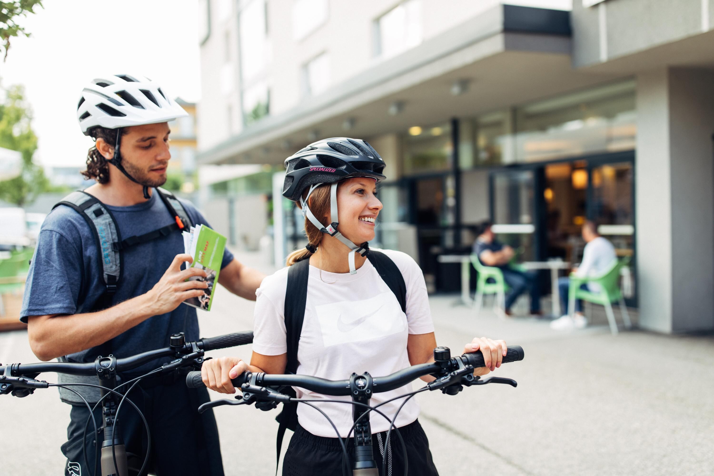 Two people on bicycles, one holding a map, in front of a modern building with outdoor seating.