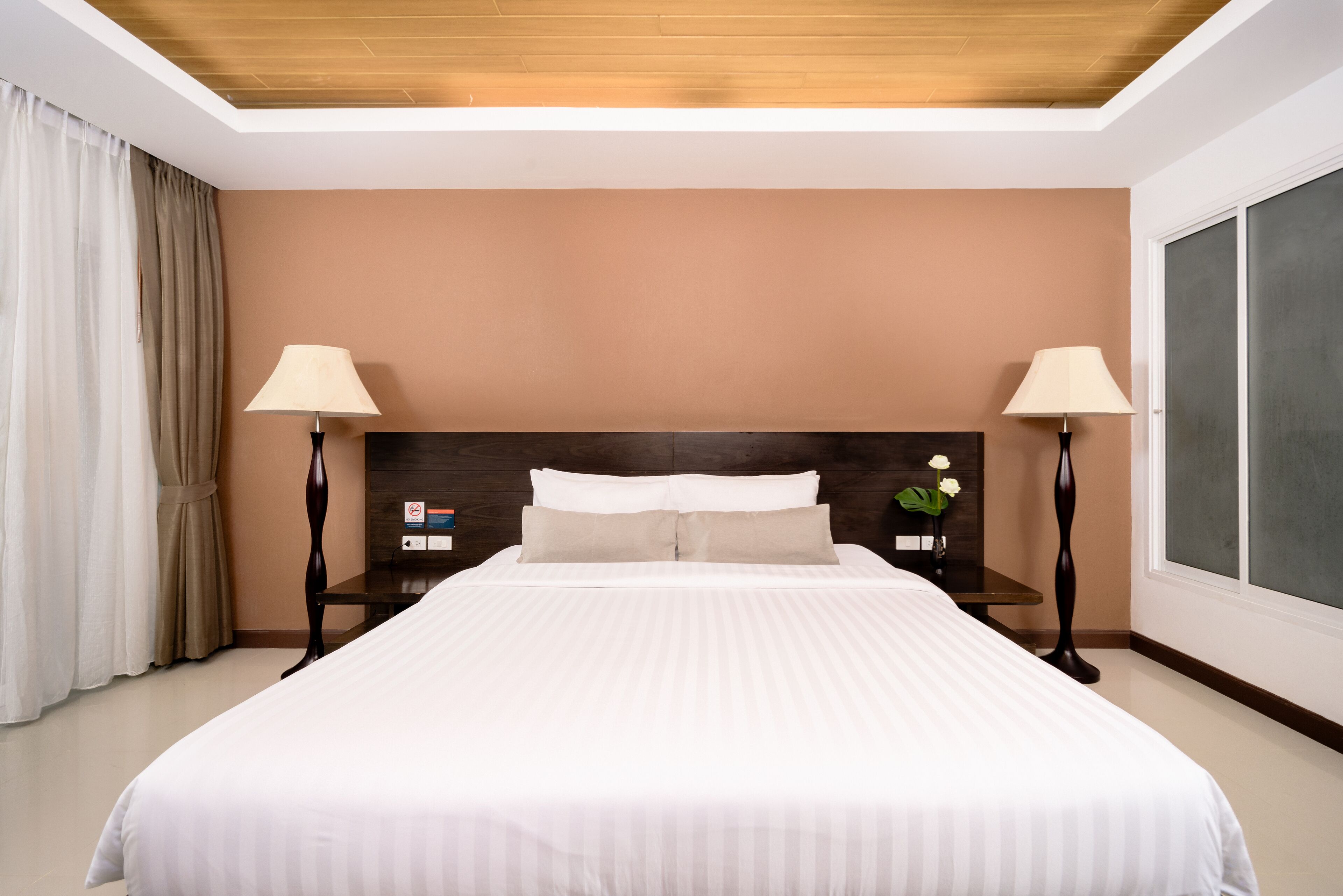 Ein Hotelzimmer mit einem großen Bett, zwei Nachttischen mit Lampen und einem Nachttisch mit einer Blume, sowie einem großen Fenster.