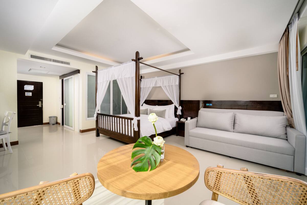 A modern hotel room with a bed, a sofa, a round table with a flower, and a chair.