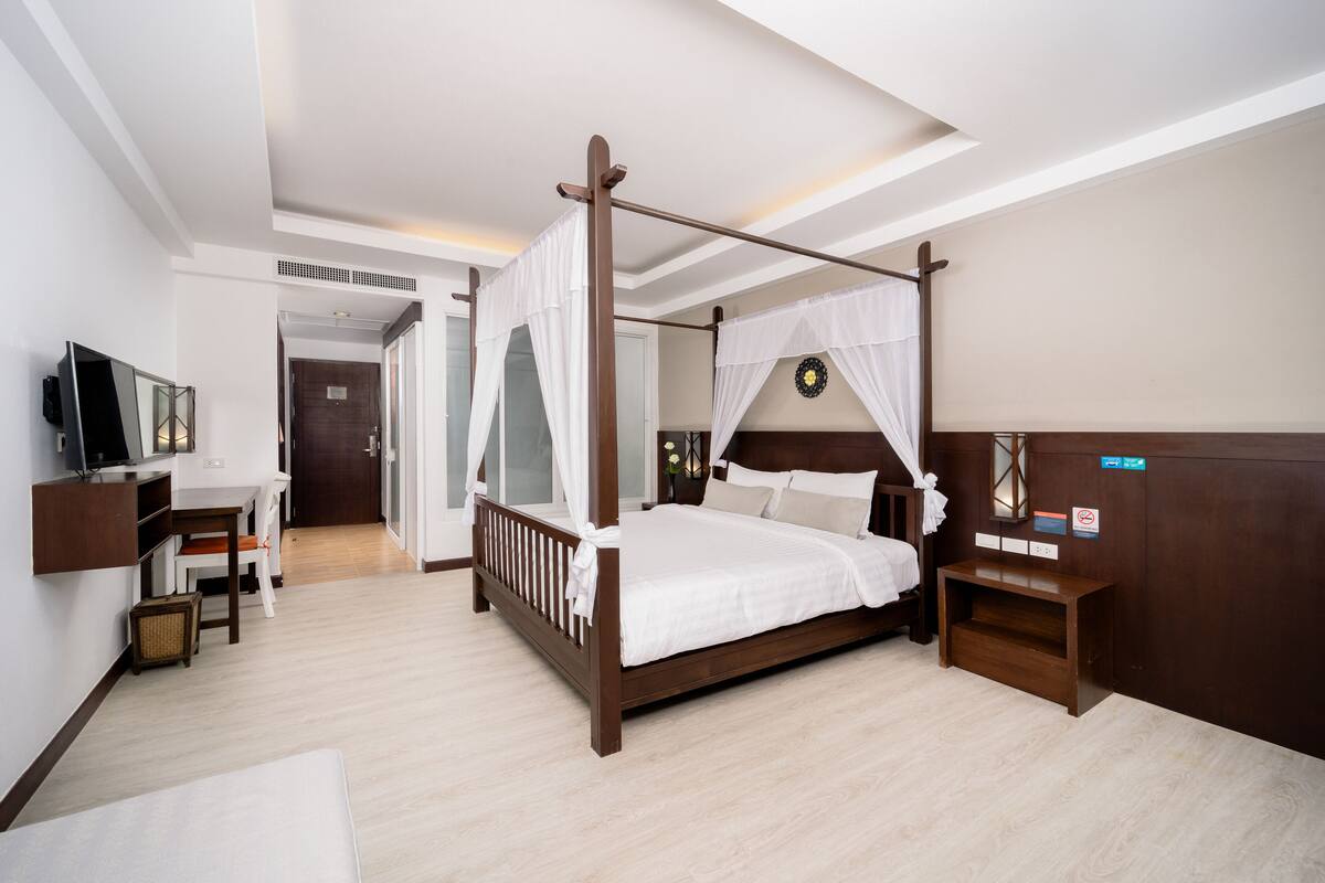 A four-poster bed with a canopy, a bedside table, a TV mounted on the wall, and a small seating area with a chair and a side table.