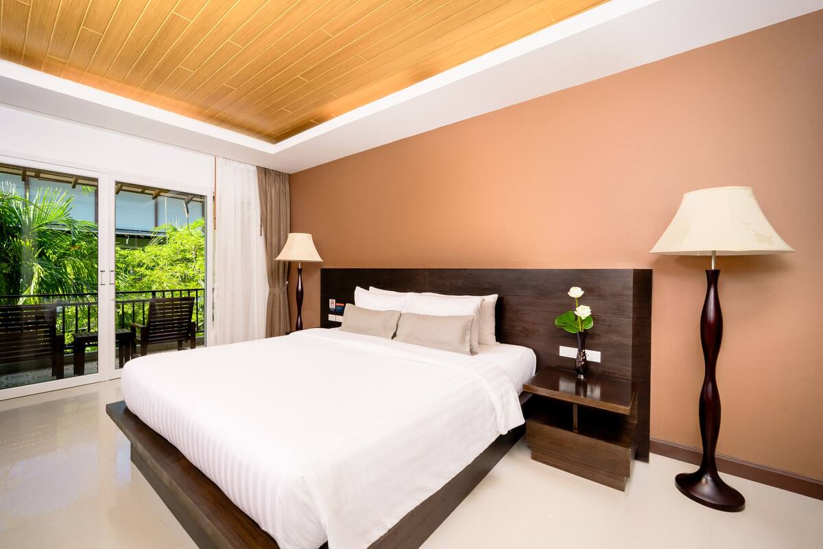 A modern hotel room with a large bed, wooden headboard, and a view of greenery through large windows.