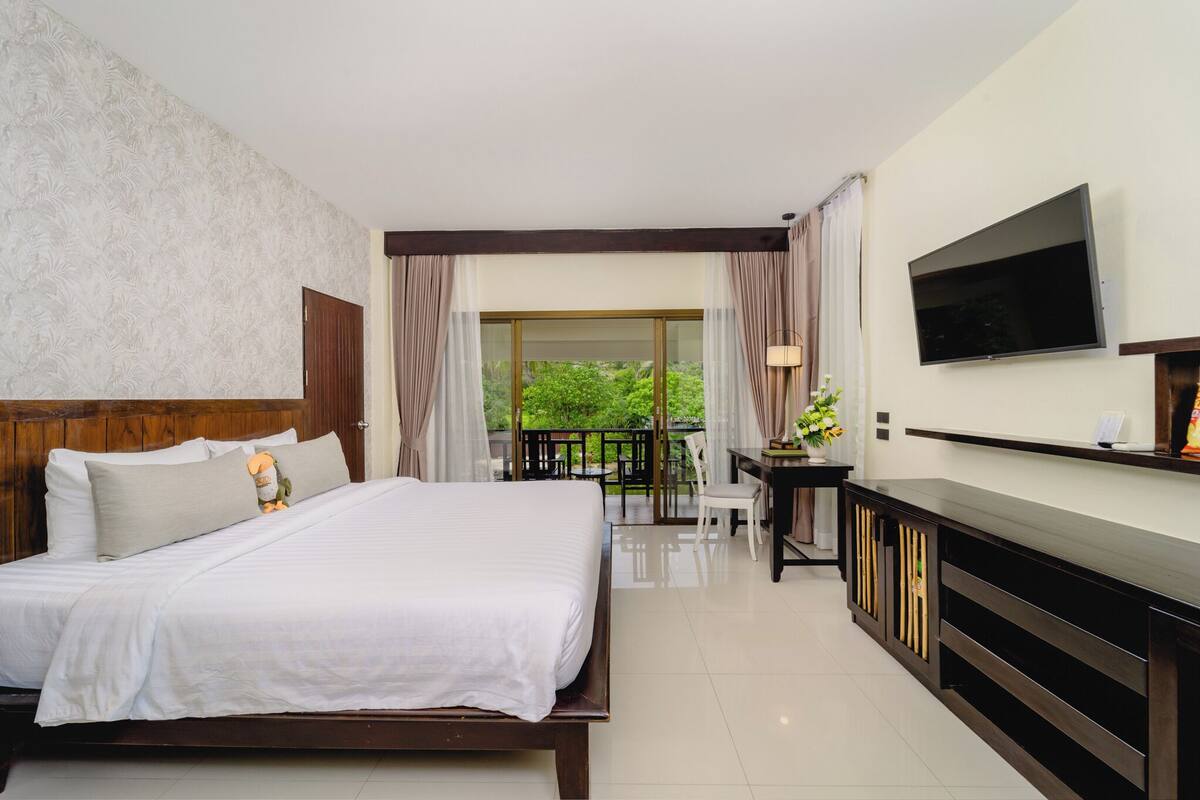 A hotel room with a bed, a TV, a desk, and a balcony with a view of greenery.