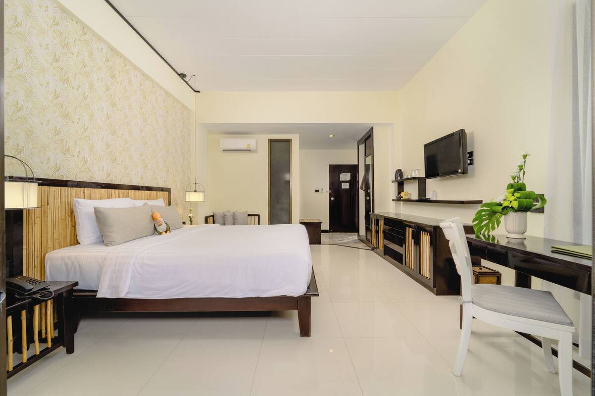 A hotel room with a bed, bedside table, lamp, TV, desk, chair, and a potted plant.