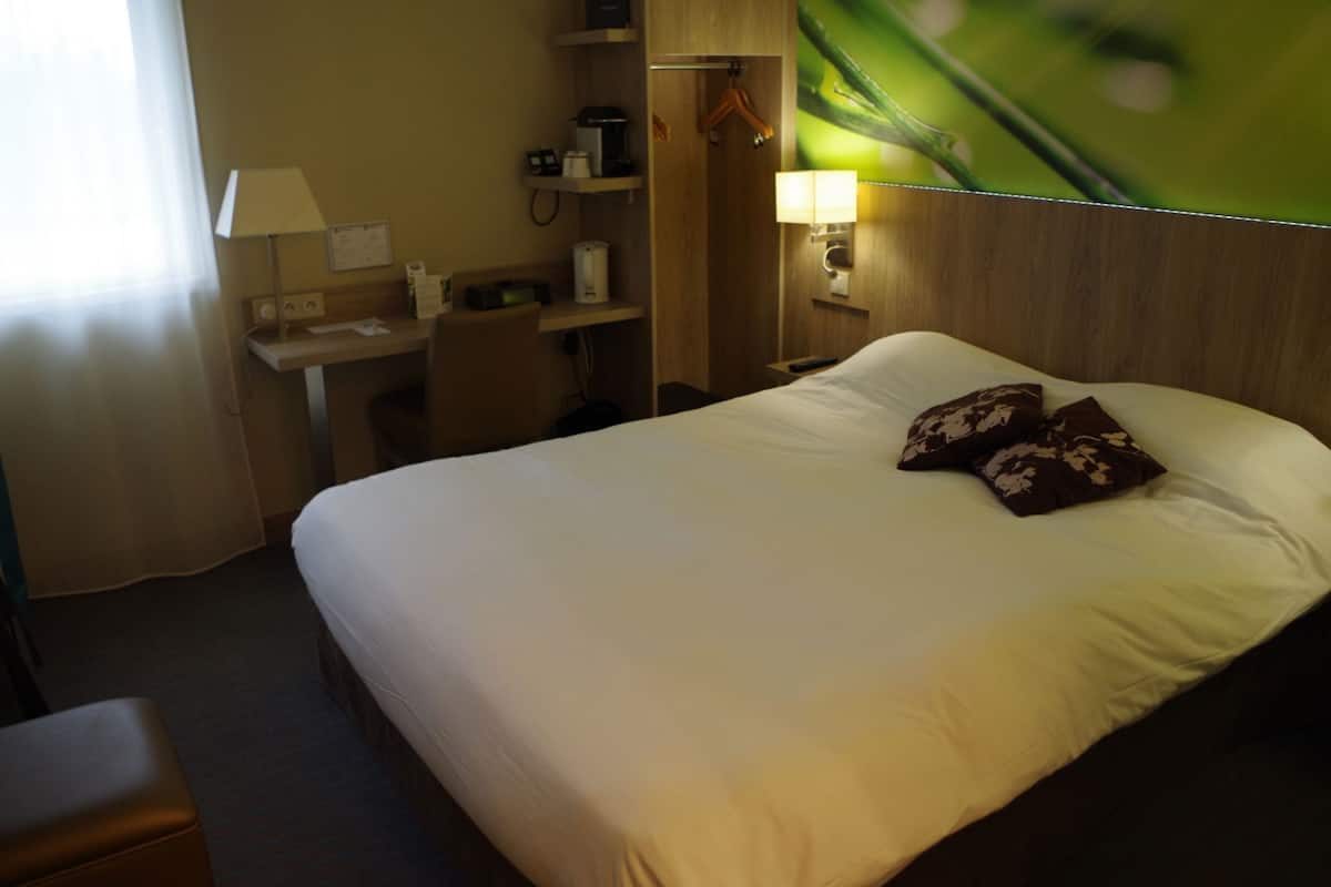 A hotel room with a bed, a desk, a chair, a lamp, and a wall-mounted shelf with a telephone and a coffee maker.