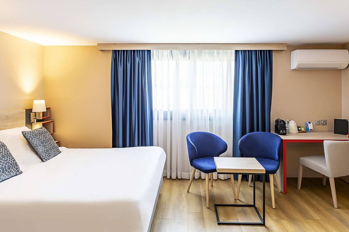 A hotel room with a bed, two blue chairs, a small desk, a white lamp, and an air conditioning unit.