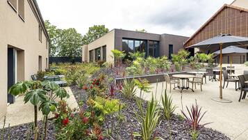 A modern outdoor patio with a variety of plants, a seating area with tables and chairs, and a building with large windows.