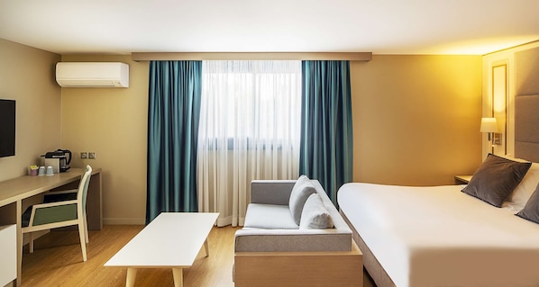 Una habitación de hotel con cama, escritorio, silla, mesita y ventana con cortinas.