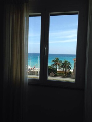 A room with a large window overlooking a beach with palm trees and the ocean. - Residence Le Saline (Borgio Verezzi)