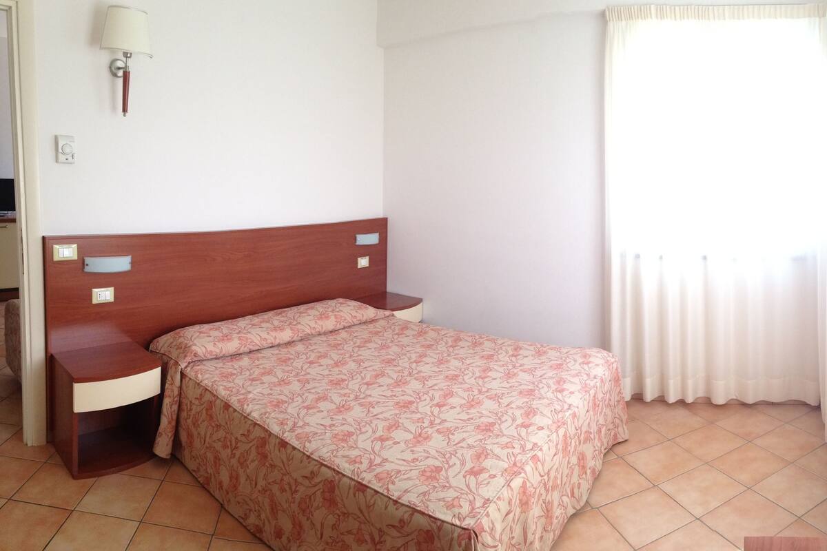 Une chambre à coucher avec une tête de lit en bois, une table de chevet, une lampe, un lit recouvert d’une couvre-lit à motifs floraux et une fenêtre avec des rideaux.