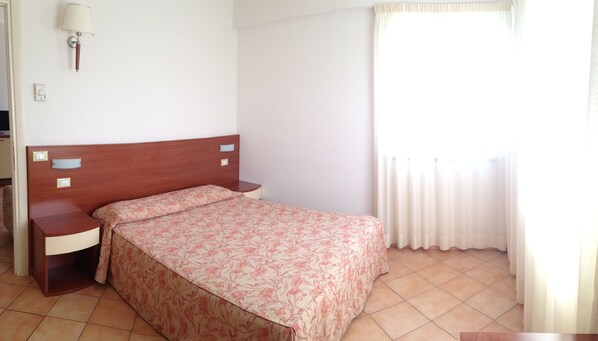 Quarto com cabeceira de madeira, mesa de cabeceira, luminária, cama com colcha florida e janela com cortinas.