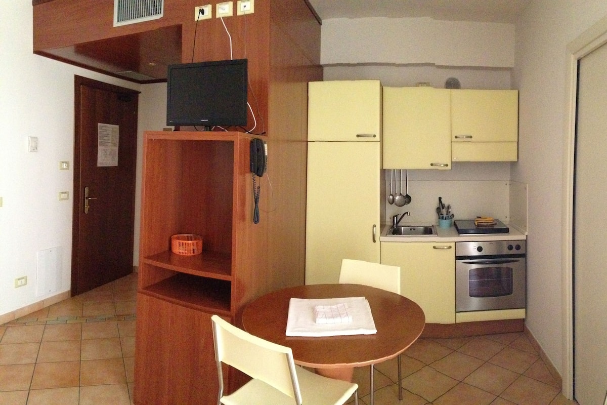 Une cuisine compacte avec un coin repas, comprenant un meuble en bois, un petit réfrigérateur, une cuisinière et un téléviseur posé sur une étagère.