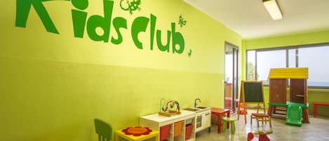 Un área de juegos infantiles con un letrero que dice 'Kids Club', una cocina de juguete, un caballo balancín y una pelota.