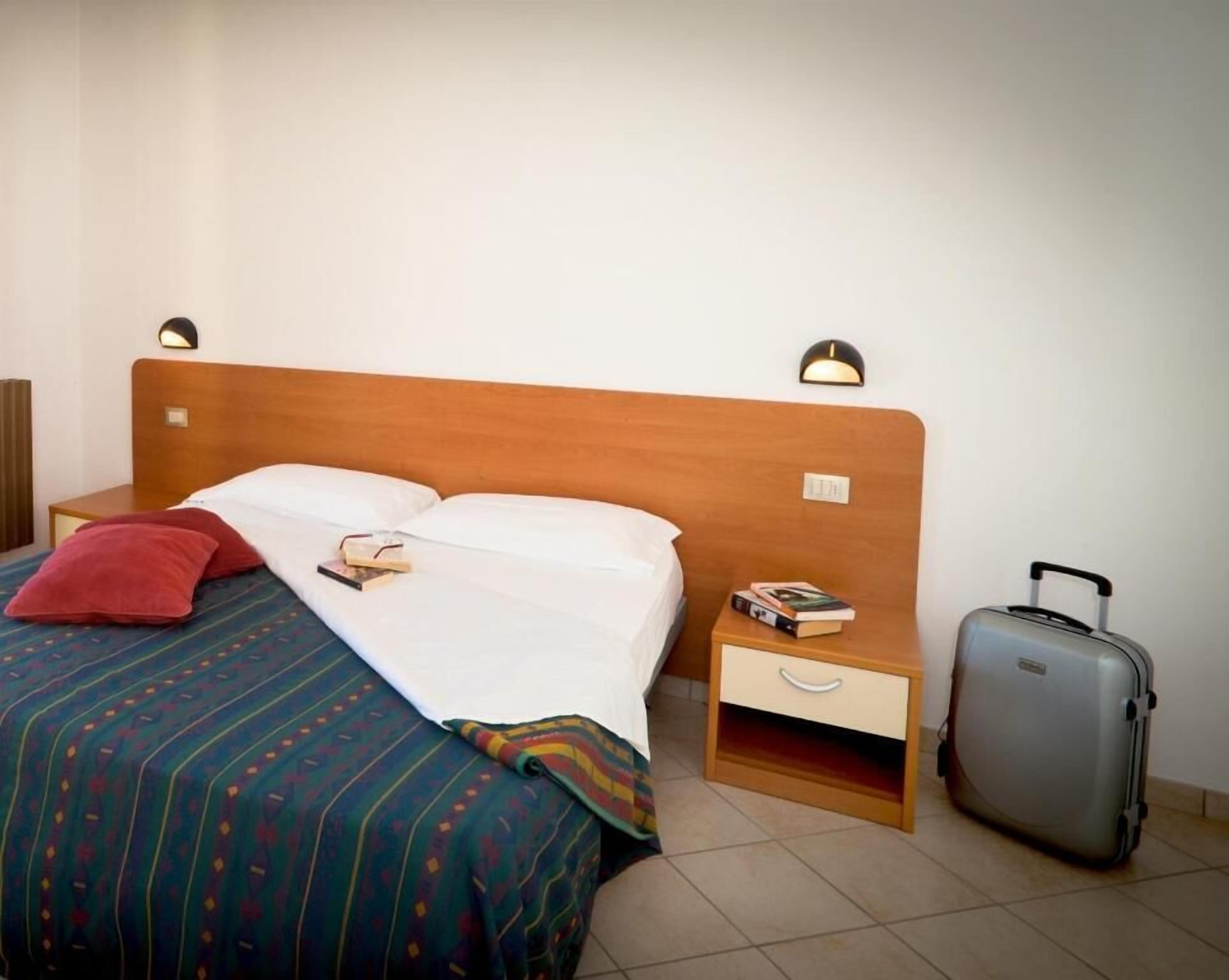 A hotel room with a wooden bed, a nightstand with a lamp, a drawer, and a suitcase.