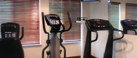Un gimnasio con máquinas de cardio, un televisor en la pared y grandes ventanas con persianas.