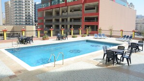 Ein Pool auf dem Dach mit Liegestühlen und Tischen, umgeben von einem Hochhaus mit mehreren Balkonen.