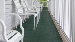 A covered porch with white railings and a green carpet, featuring rows of white chairs and a visible exit sign.