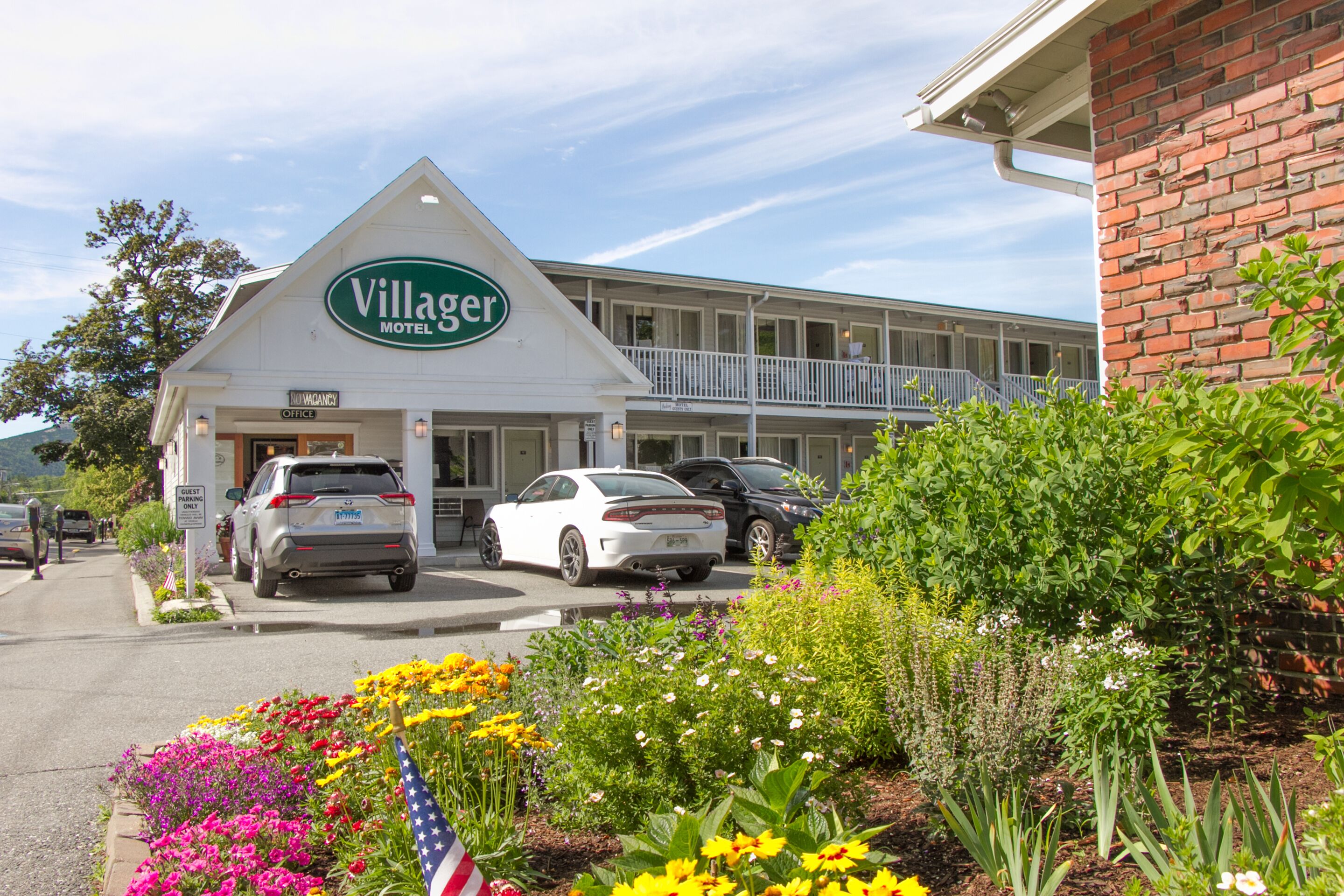 A motel with a prominent sign, a parking lot with cars, and a landscaped garden with flowers.