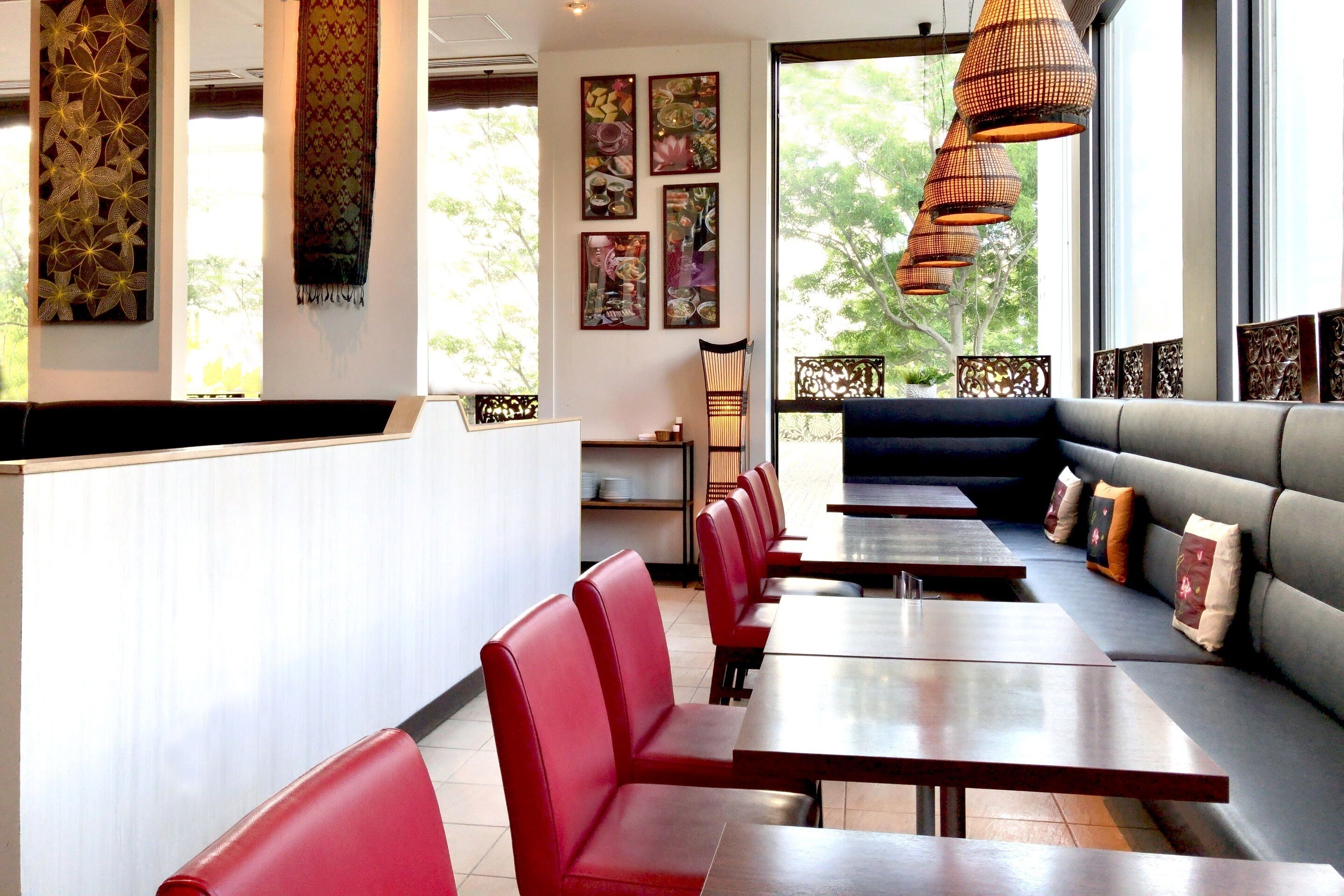 A restaurant with red chairs, wooden tables, and a grey sofa. There are decorative wall hangings and a large window with a view of trees.