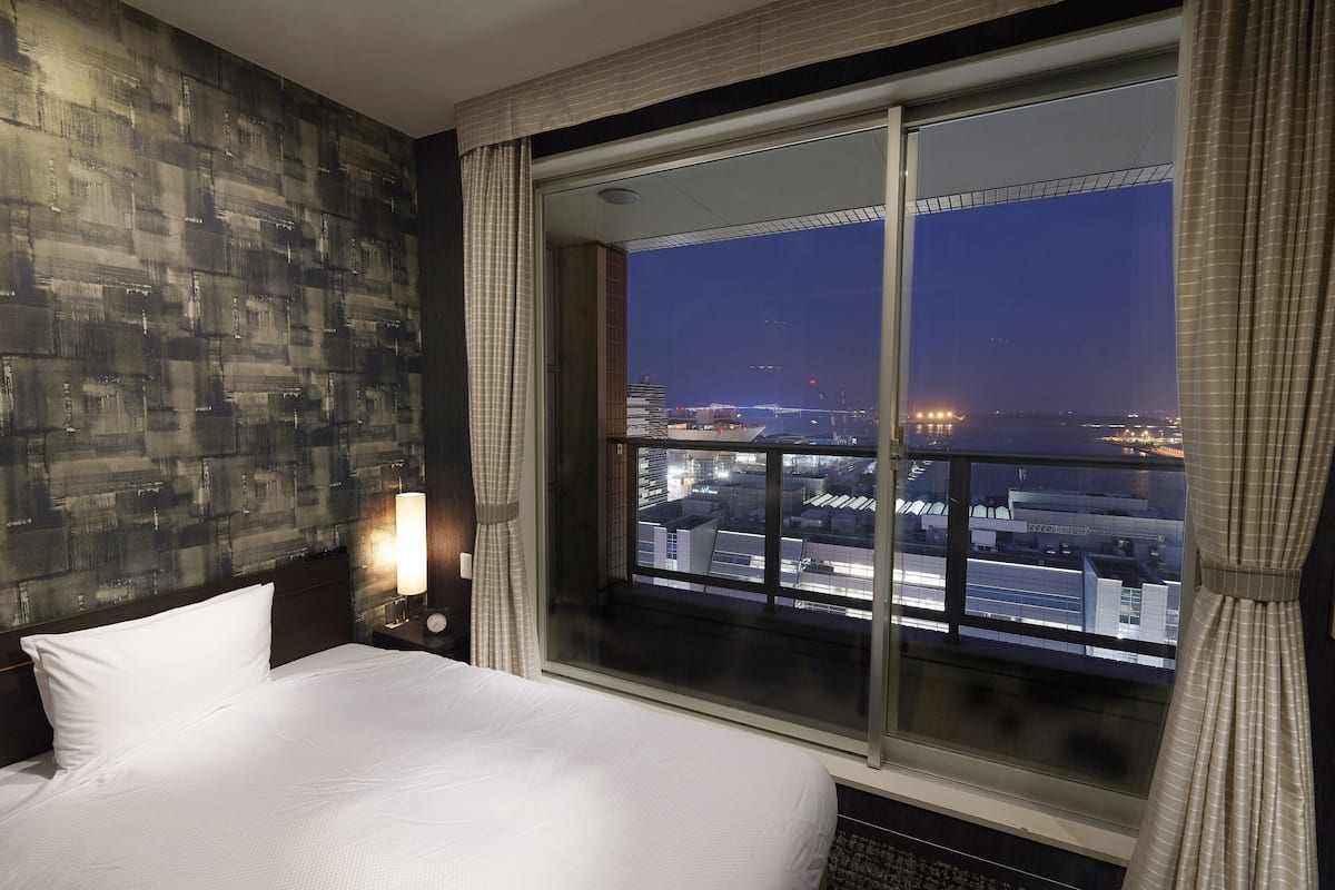 A hotel room with a large bed, a nightstand with a lamp, and a view of a cityscape through a large window.