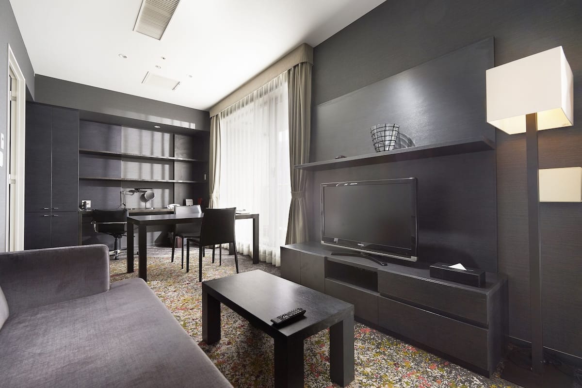 A modern hotel room with a dark color scheme, a flat-screen TV, a sofa, a coffee table, a desk with a chair, and a large window with curtains.