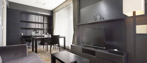 A modern hotel room with a dark color scheme, a flat-screen TV, a sofa, a coffee table, a desk with a chair, and a large window with curtains.