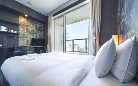 A hotel room with a large bed, a flat-screen TV, a desk with a chair, and a view of the city through the window.