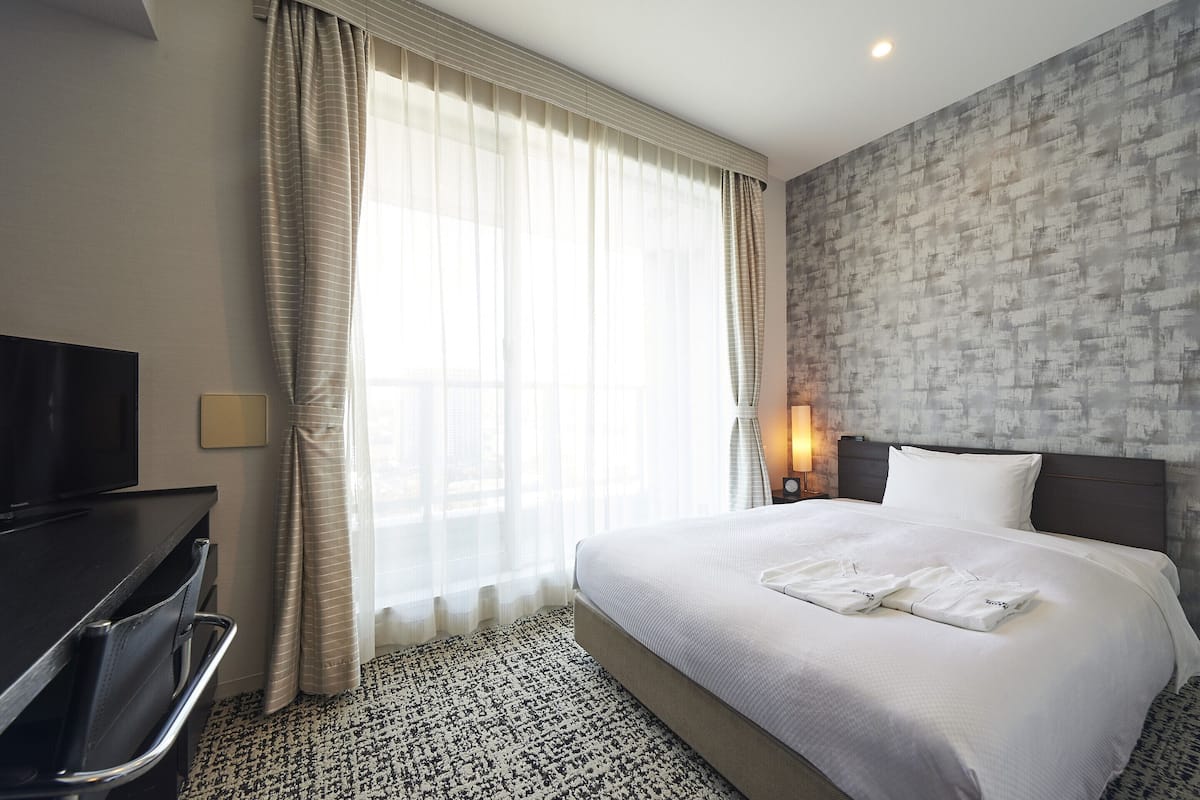A hotel room with a large bed, a flat-screen TV, a desk, and a window with sheer curtains.