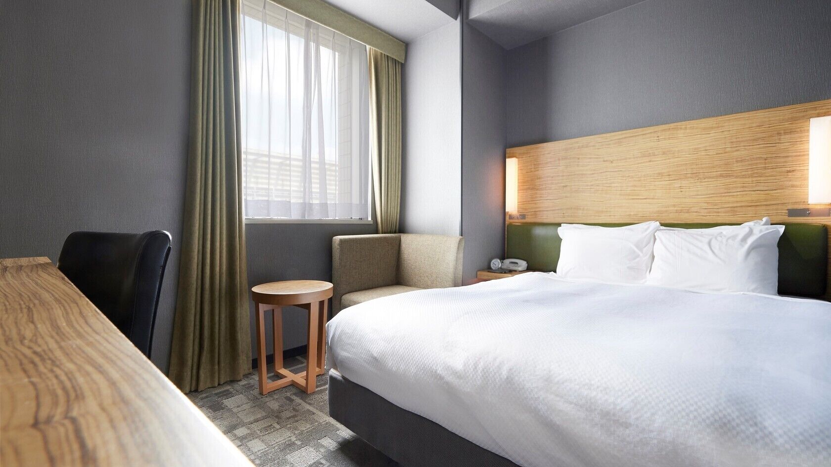 A hotel room with a bed, a chair, a small table, and a window with curtains.