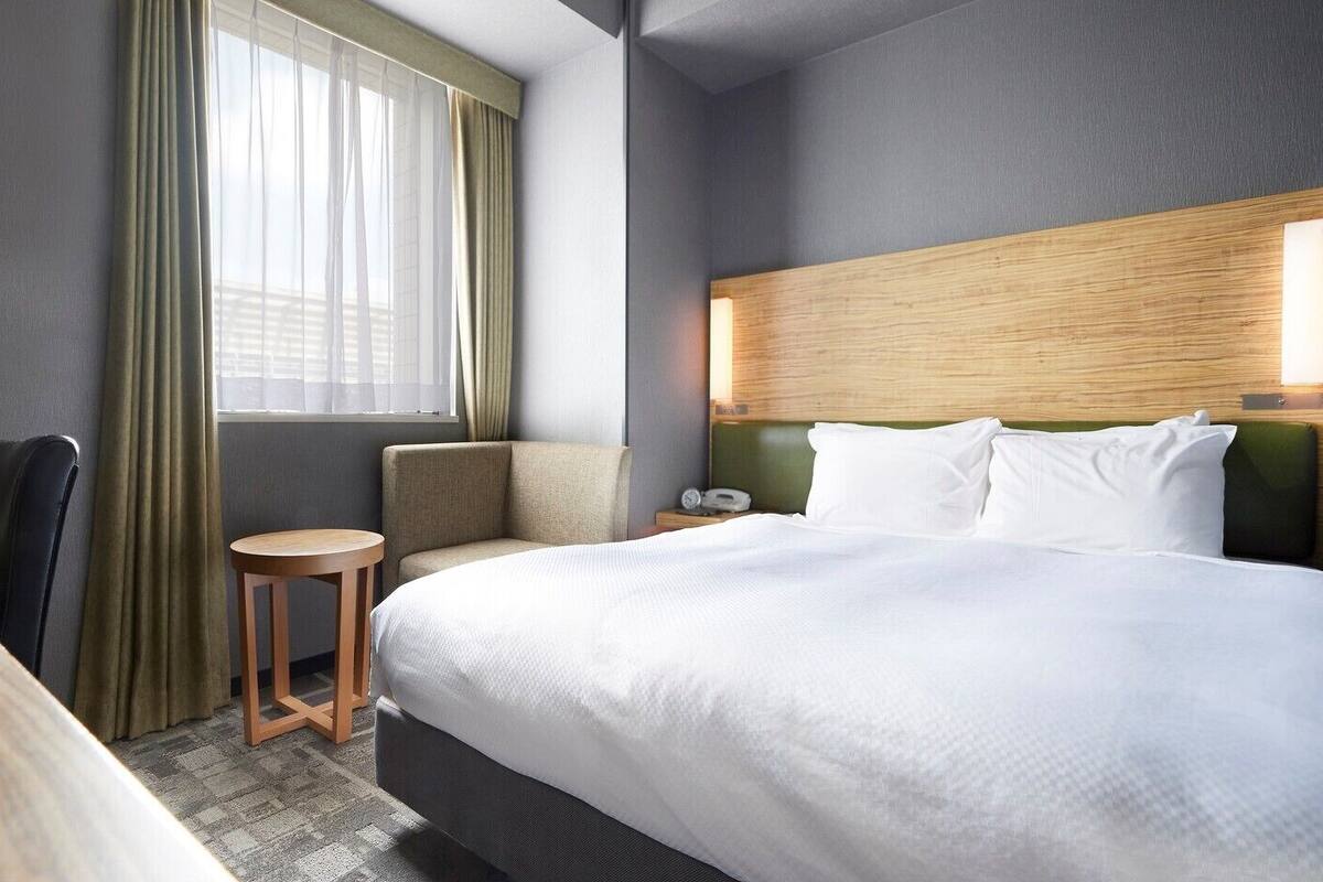 A hotel room with a bed, a chair, a small table, and a window with curtains.
