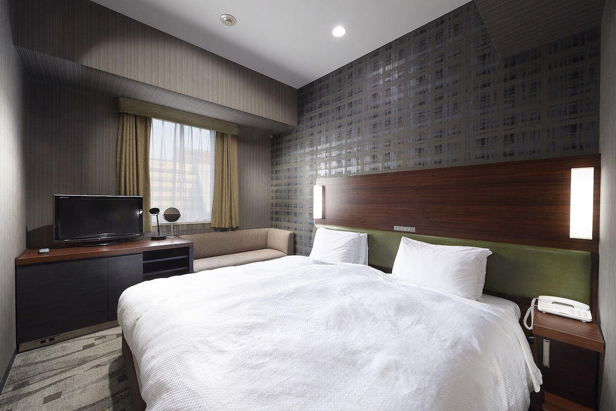 A hotel room with a large bed, a TV on a wooden console, a bedside phone, and a window with curtains.