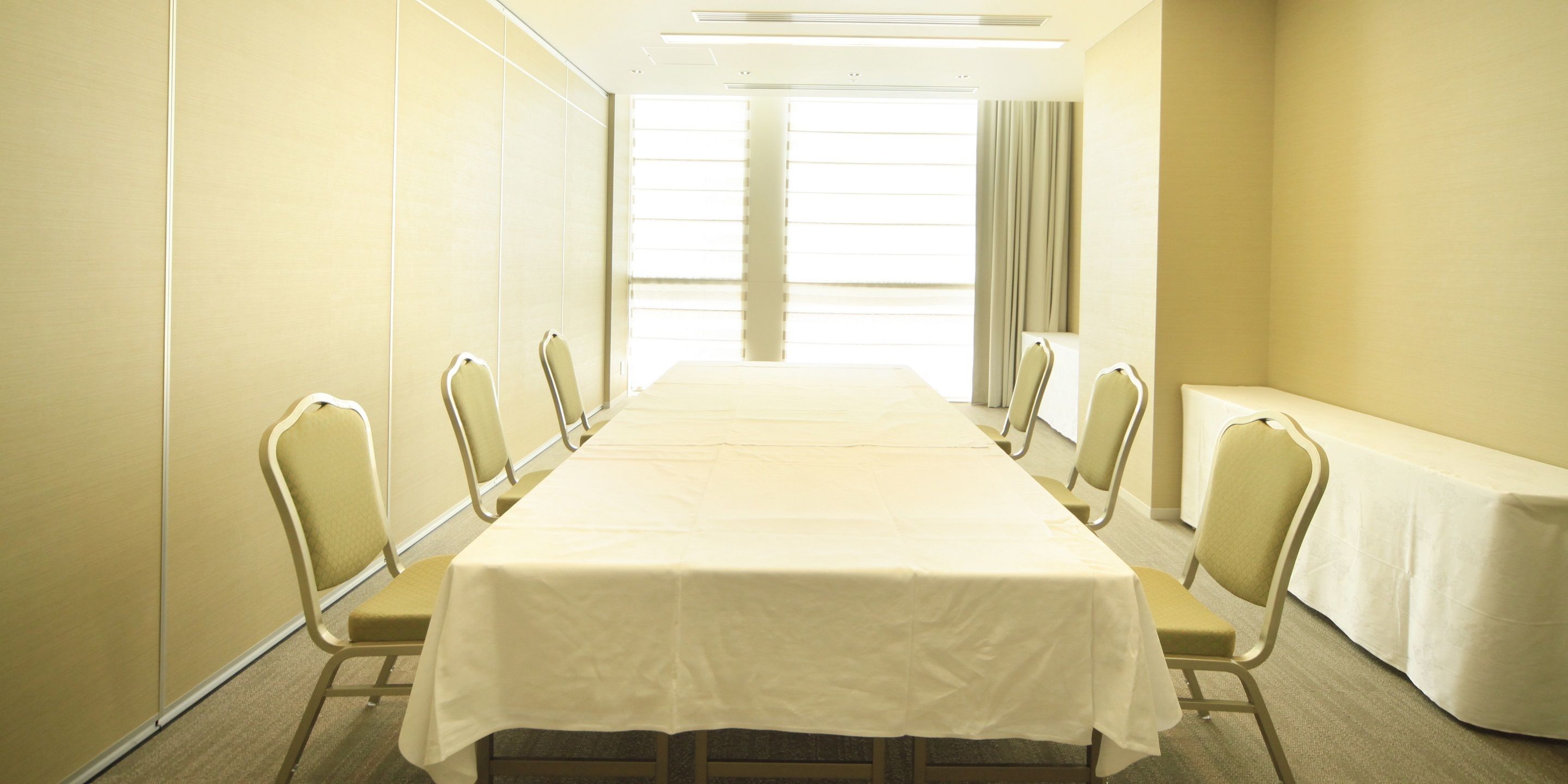 Un salón de conferencias con una larga mesa y sillas, ventanas amplias con persianas y piso alfombrado.