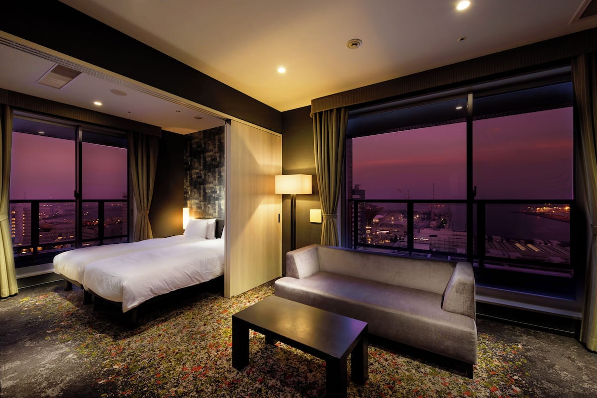 A hotel room with a bed, a sofa, a coffee table, and large windows offering a city view.