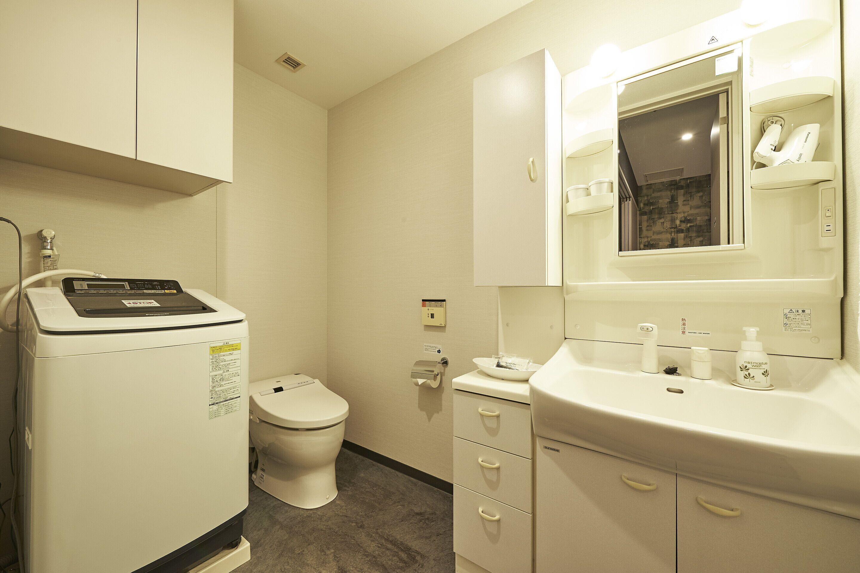A bathroom with a white toilet, a washing machine, a sink with toiletries, and a mirror.