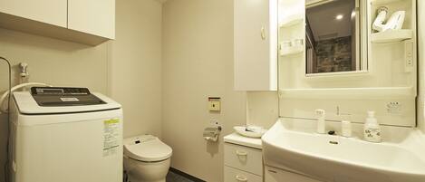 A bathroom with a white toilet, a washing machine, a sink with toiletries, and a mirror.