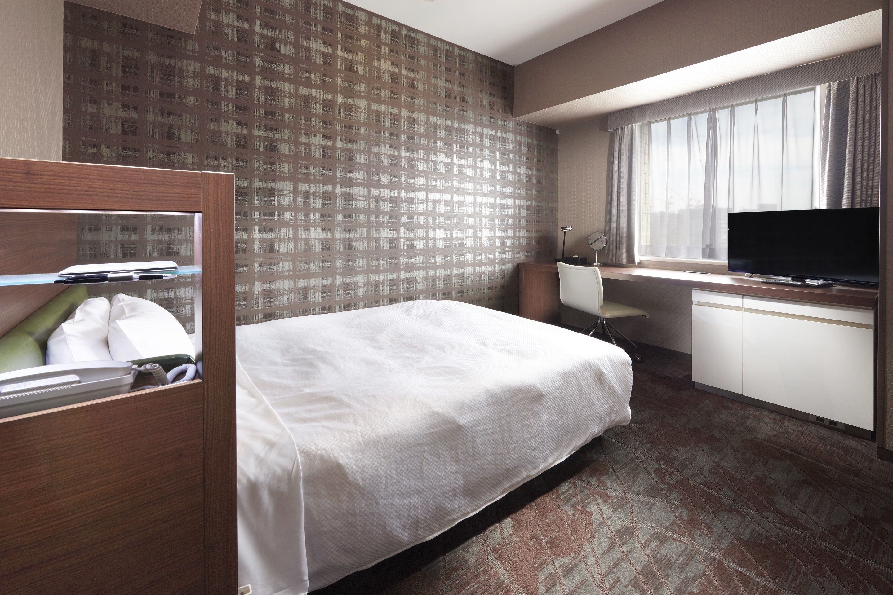 A hotel room with a bed, desk, TV, and a window with curtains.