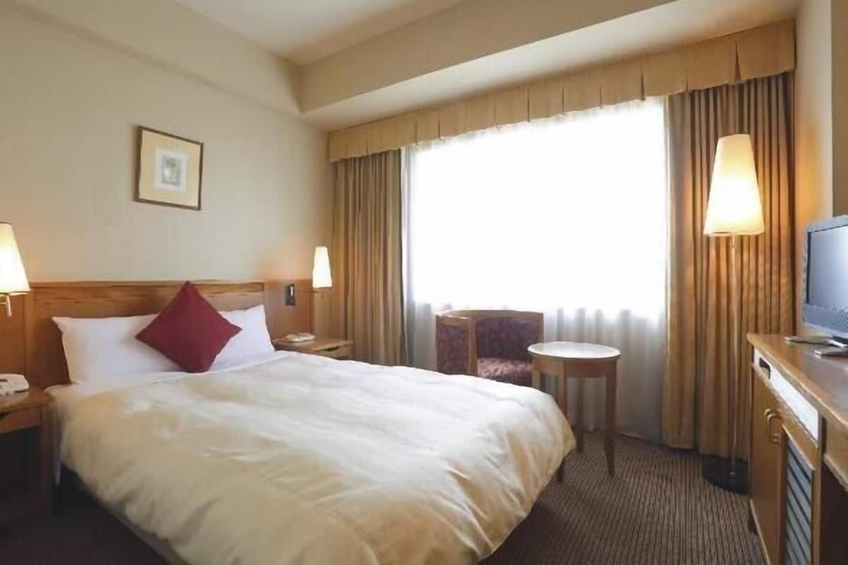 Una habitació d'hotel amb un llit, una cadira, una taula petita, una televisió i cortines.