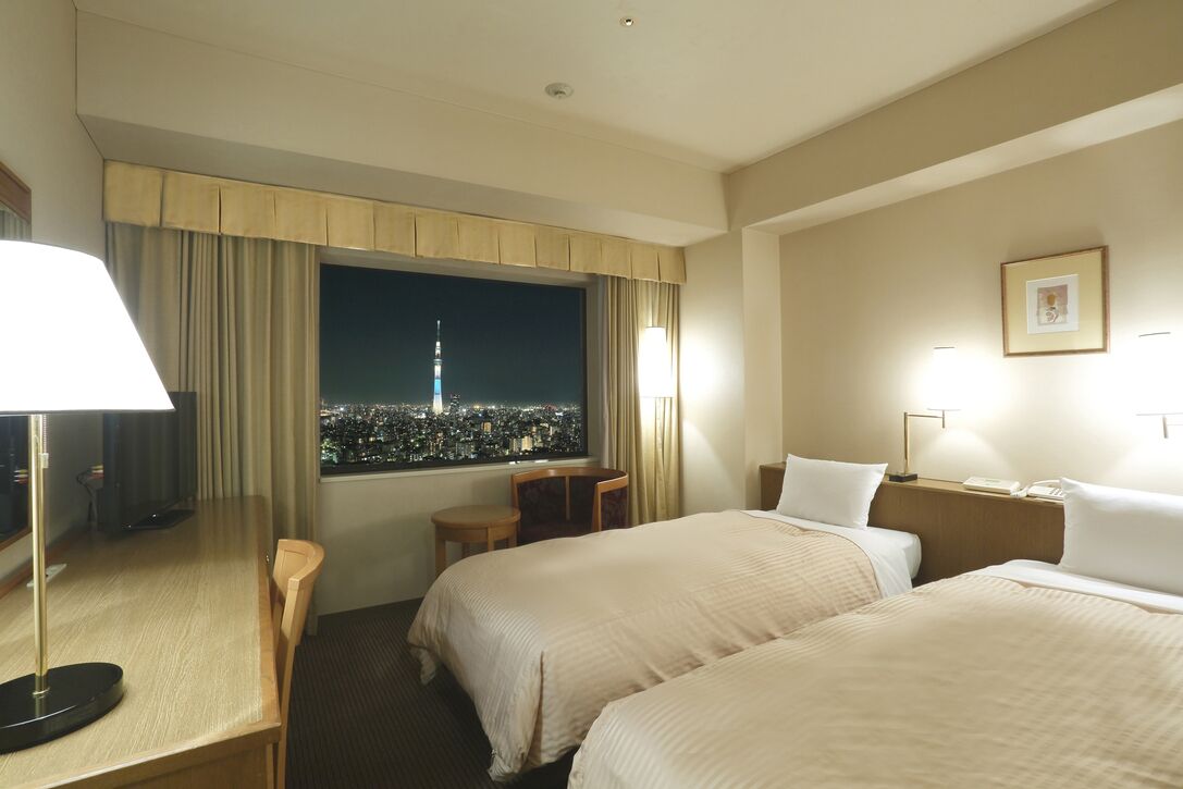 A hotel room with two beds, a desk, a chair, a lamp, a window with a cityscape view, and a framed picture on the wall.