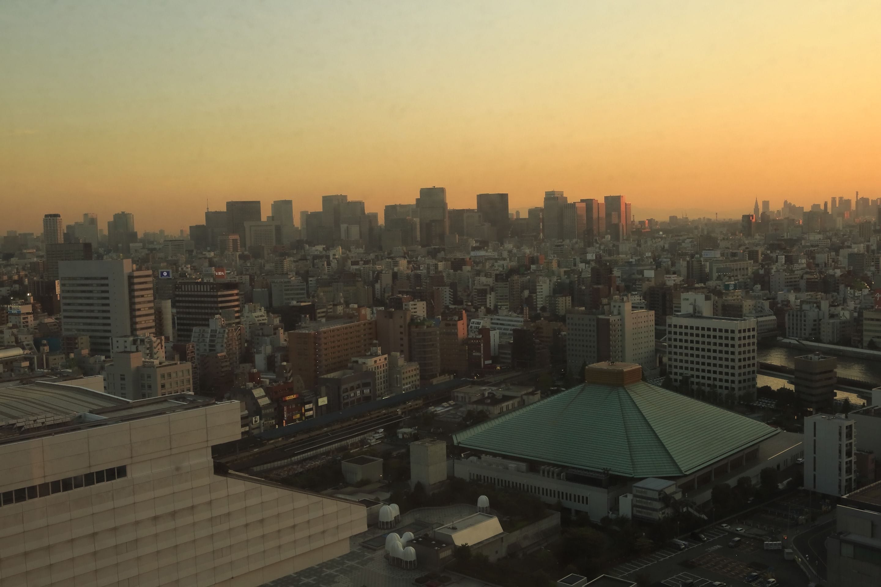An aerial view of a cityscape with a mix of high-rise buildings and a prominent green-roofed structure in the foreground.