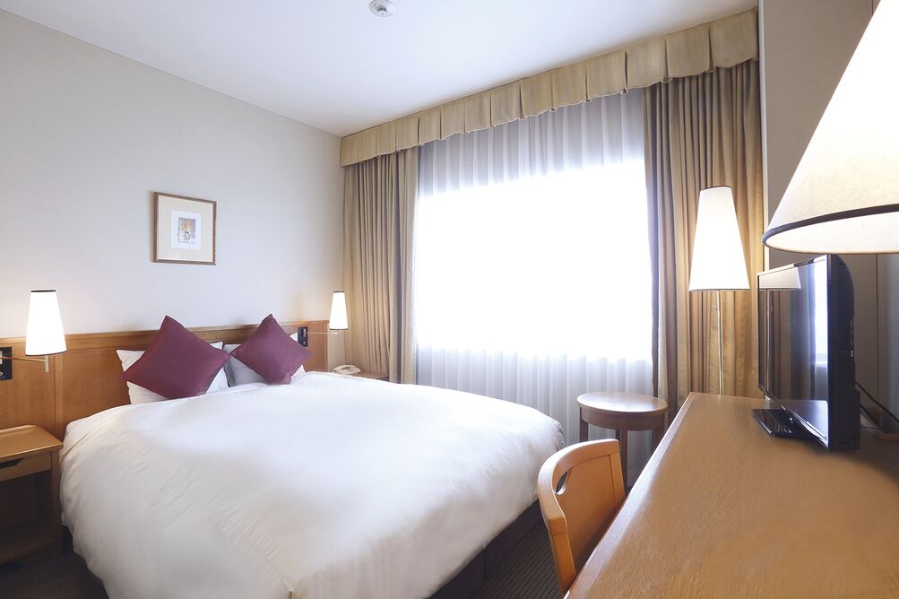 A hotel room with a large bed, two bedside lamps, a desk with a chair, a television, and a window with curtains.