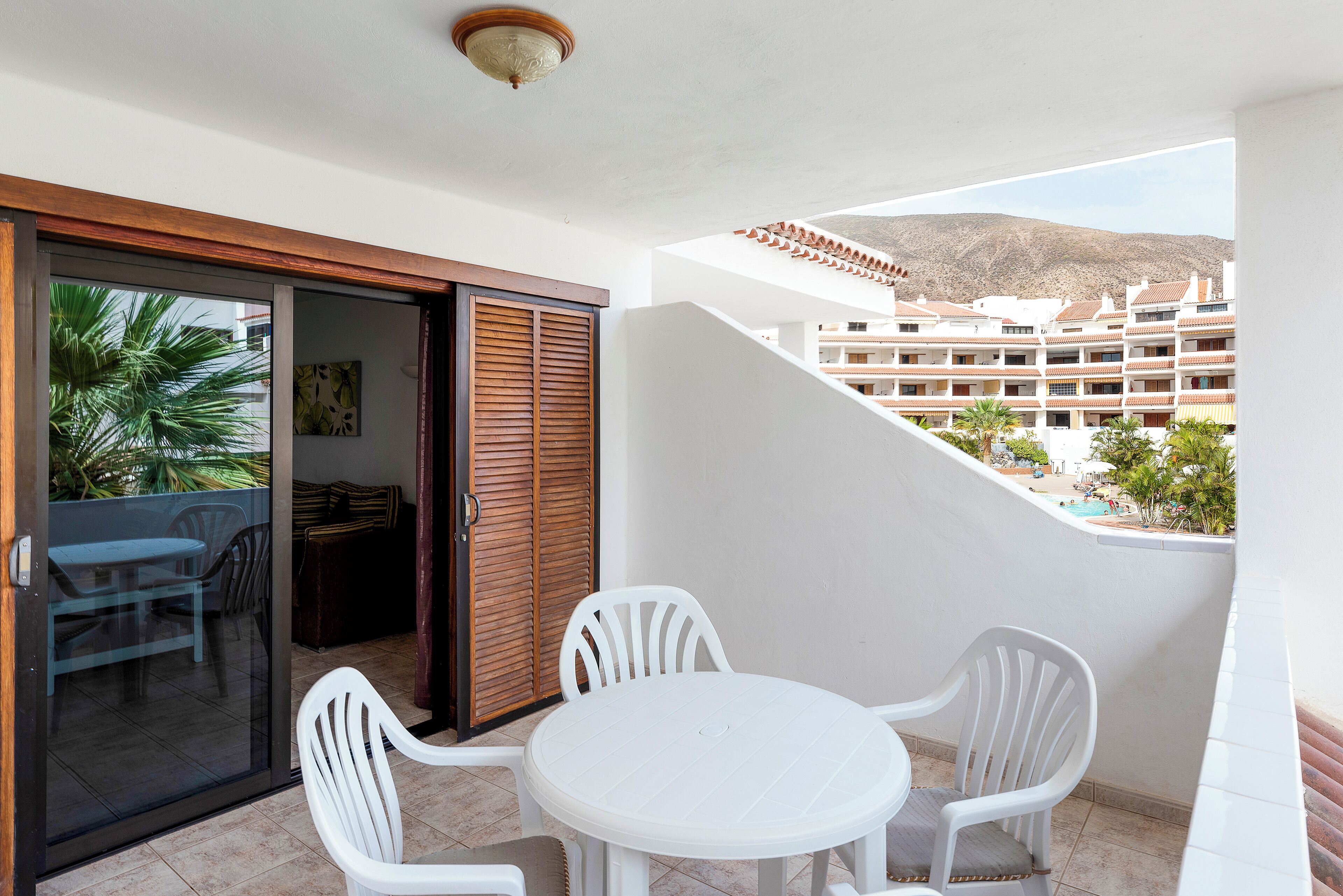 A balcony with a white table and chairs, a sliding glass door, and a view of a resort with buildings and mountains in the background.