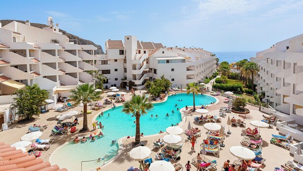 A resort with a central swimming pool surrounded by sun loungers and umbrellas, multiple floors of white buildings, and a clear view of the sea. - Paloma Beach Apartments (Arona)