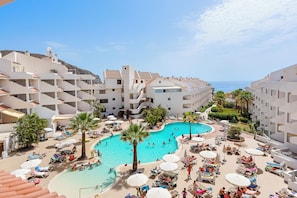 A resort with a central swimming pool surrounded by sun loungers and umbrellas, multiple floors of white buildings, and a clear view of the sea. - Paloma Beach Apartments (Arona)