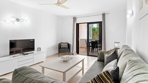 A modern living room with a flat-screen TV, a ceiling fan, a sofa, and a dining area visible through a glass door. - Paloma Beach Apartments (Arona)