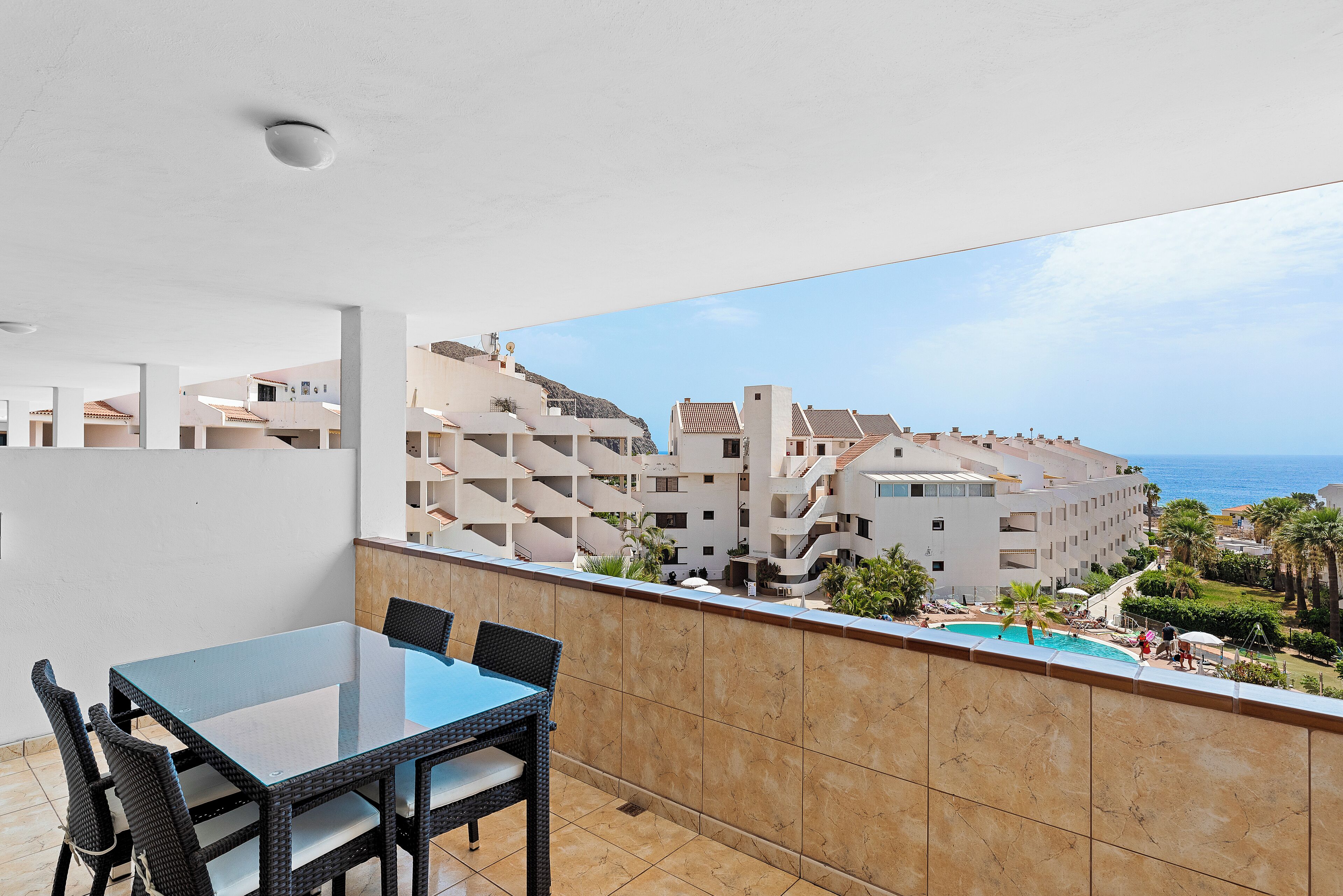 Un balcón con mesa y sillas de vidrio, con vista a una piscina y una zona residencial con varios edificios.