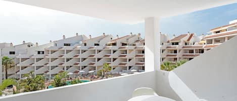 A balcony with white chairs and a table, overlooking a pool area with umbrellas and palm trees.
