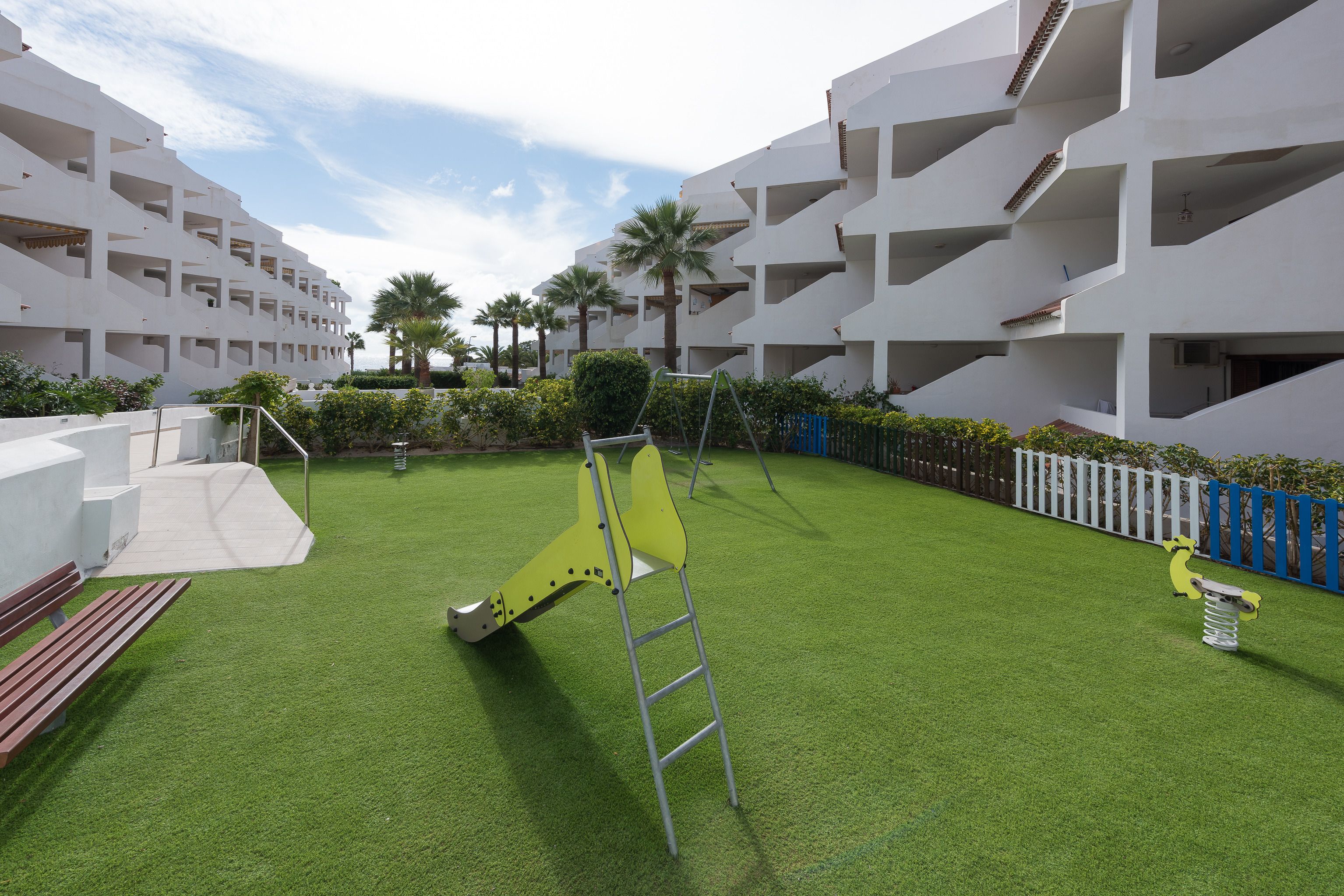 A courtyard with artificial grass, a yellow swing set, a ladder, and a white fence.