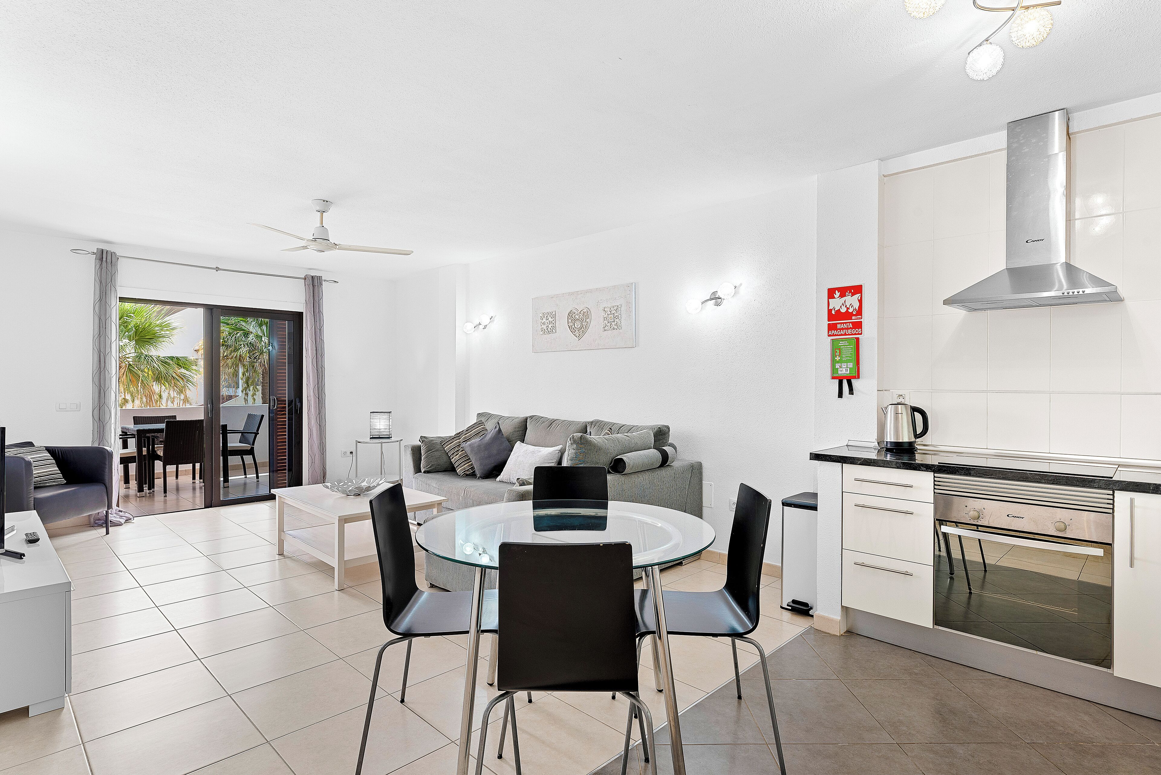 Una cucina moderna con forno integrato, una zona pranzo con tavolo in vetro e sedie nere, e un divano grigio in un soggiorno con ventilatore a soffitto.