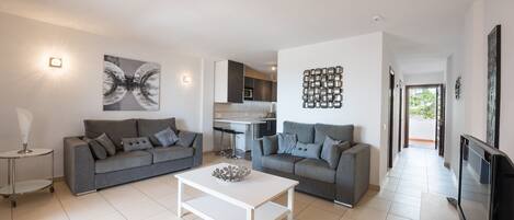 A modern living room with two grey sofas, a white coffee table, and a kitchen in the background.