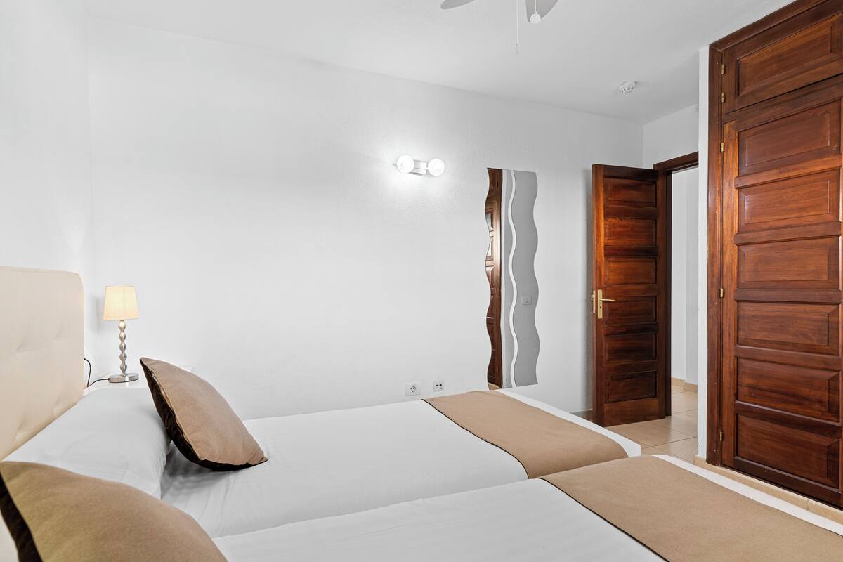 A bedroom with two beds, a nightstand, a lamp, a wall-mounted light, and a wooden door.
