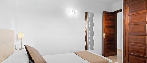 A bedroom with two beds, a nightstand, a lamp, a wall-mounted light, and a wooden door.